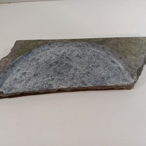 May include: A broken piece of gray and green stone with a rounded, indented section. The stone has a rough, uneven texture.