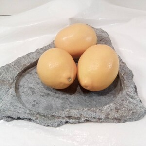 May include: Three yellow lemons on a gray stone serving dish. The dish is shaped like a heart and has a raised rim.