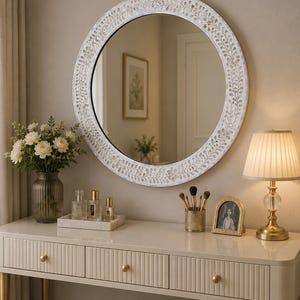 May include: A round wall mirror featuring a white frame with an intricate, carved floral and vine pattern. The mirror is positioned above a cream-colored vanity table with three drawers, gold-toned knobs, and slender gold-toned legs.