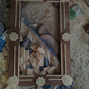 May include: A rectangular wooden frame featuring light-colored, carved floral accents at each corner and along the center of the sides. The frame is constructed with inlaid straight pieces and circular flower motifs.