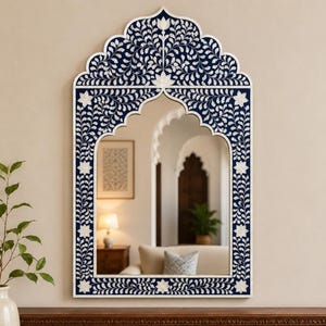 May include: A rectangular wall mirror with an arched top, featuring a dark blue frame inlaid with white floral and vine patterns. The frame has a scalloped edge design, and the reflective glass surface shows a room interior with a lamp and furniture.