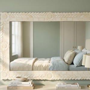 May include: A rectangular wall mirror featuring a wide, off-white frame with intricate carved floral and leaf patterns. The frame displays circular motifs at each corner and center, surrounding a clear glass surface reflecting a bedroom interior.