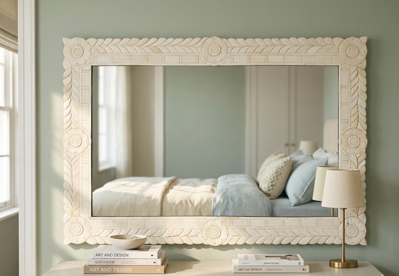 May include: A rectangular wall mirror featuring a wide, off-white frame with intricate carved floral and leaf patterns. The frame displays circular motifs at each corner and center, surrounding a clear glass surface reflecting a bedroom interior.