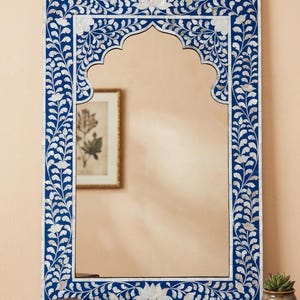 May include: Rectangular wall mirror with a blue and white floral design. The frame features a dark blue background with white floral and leaf patterns. The mirror has an arched top and is hanging from a gold chain.