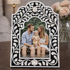 May include: A rectangular picture frame with an arched top, featuring a black background with white floral and leaf inlay patterns. The frame includes two small flower-shaped accents at the top and bottom center, each with a central pearl-like detail.