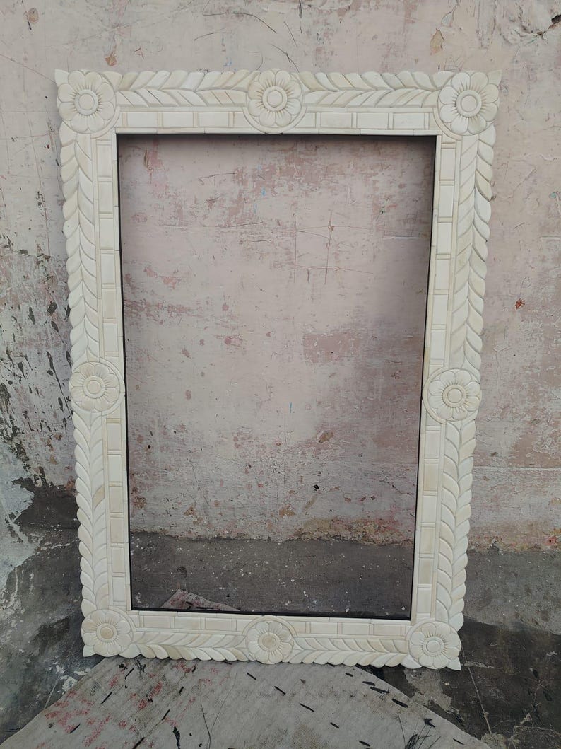 May include: A rectangular mirror frame featuring an off-white, cream-colored finish with intricate hand-carved floral motifs at each corner and center point, complemented by a repeating leaf-like pattern along the entire border edge.