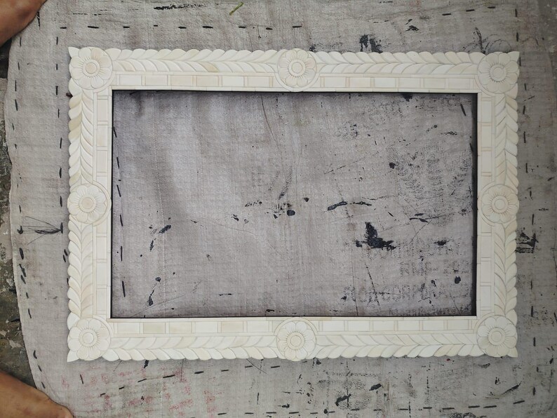 May include: A rectangular off-white picture frame featuring intricate carved leaf patterns along the borders and circular floral motifs at each corner and the center of every side, creating a detailed and symmetrical decorative border.