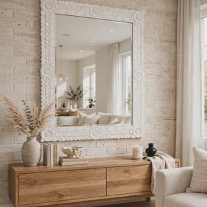 May include: A large rectangular wall mirror with a thick white frame featuring carved floral and leaf patterns. The mirror is mounted on a light-colored brick wall above a wooden console table with two drawers and a smooth, natural wood grain finish.