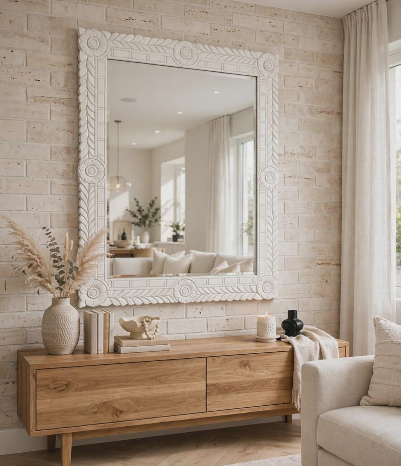 May include: A large rectangular wall mirror with a thick white frame featuring carved floral and leaf patterns. The mirror is mounted on a light-colored brick wall above a wooden console table with two drawers and a smooth, natural wood grain finish.