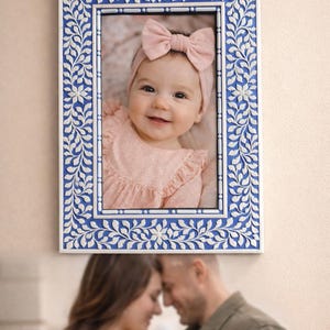 Könnte beinhalten: Ein rechteckiger Bilderrahmen mit blauem und weißem floralem Design. Der Rahmen umrahmt ein Foto eines lächelnden Babys mit einer rosa Schleife. Der Rahmen hängt an einer hellen Wand.