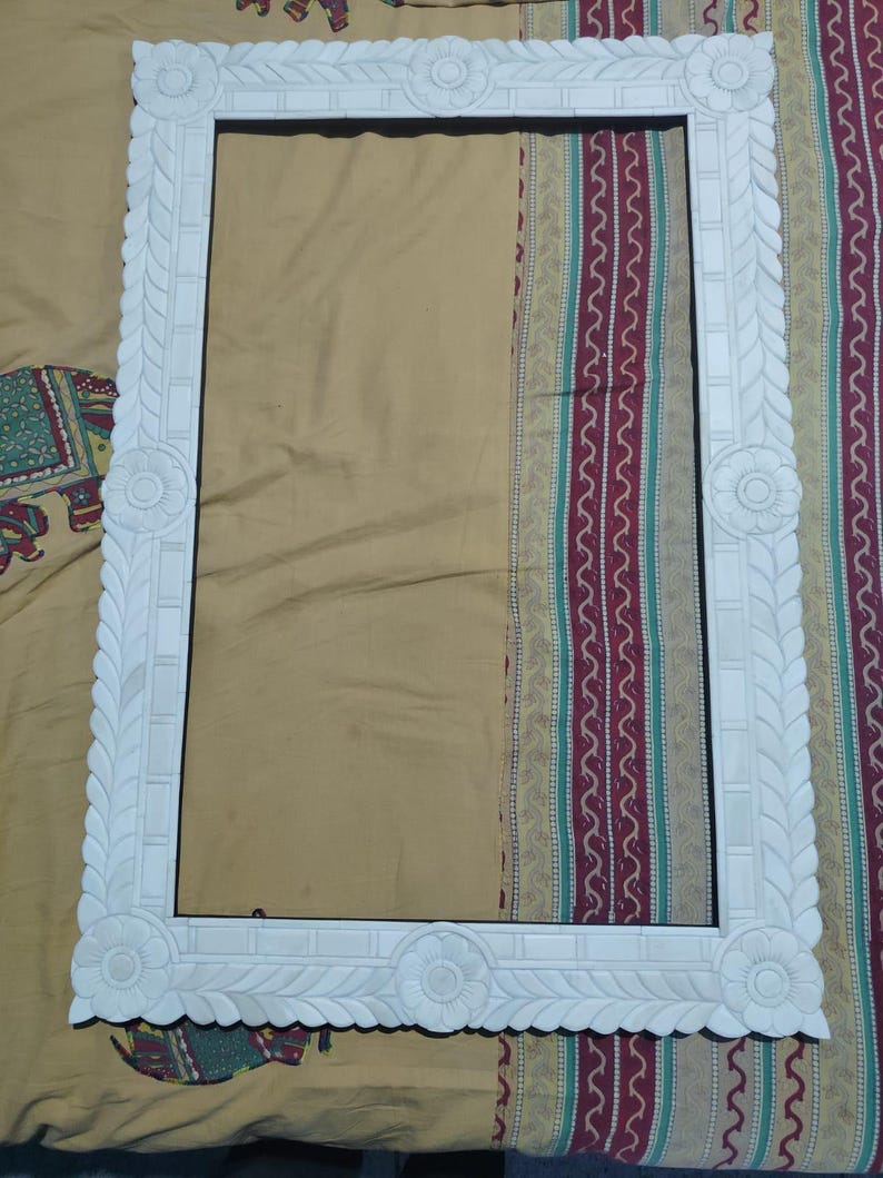 May include: A rectangular white picture frame featuring intricate carved floral motifs at each corner and along the center of each side. The frame border is decorated with a repeating leaf pattern and smooth rectangular segments.