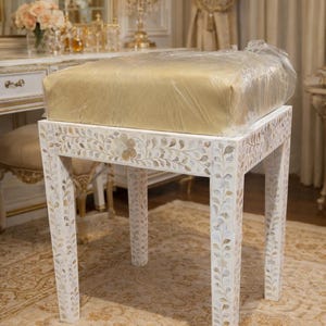 Puede incluir: Un taburete de tocador blanco con un cojín dorado cubierto de plástico. Las patas y el marco del taburete están decorados con un patrón floral. El taburete está colocado sobre una alfombra estampada.