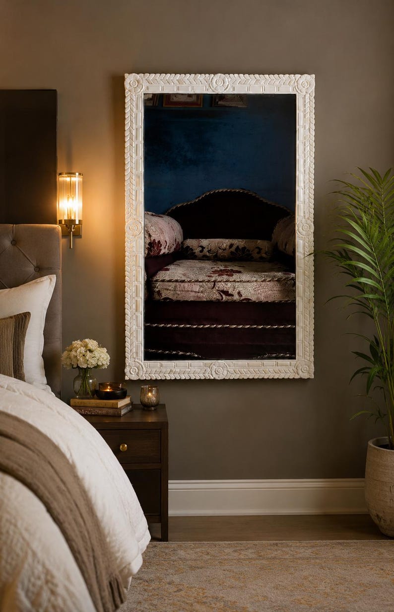 May include: A rectangular wall mirror featuring a thick, white, hand-carved frame with intricate leaf-like patterns along the edges and circular floral motifs at the center of each side, reflecting a dark room with a patterned armchair.