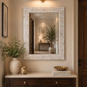 May include: A rectangular wall mirror featuring a frame with an intricate white and cream floral vine inlay pattern. The mirror is mounted above a dark wood console table with two drawers, a stone top, and a brass light fixture positioned above it.