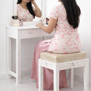 May include: A white vanity table with a rectangular mirror and a matching stool featuring intricate mother-of-pearl inlay patterns on the legs and frame. The vanity includes a single drawer with a clear crystal-style knob and a padded seat.