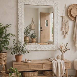 May include: A large rectangular wall mirror with a thick, off-white carved wooden frame featuring floral and leaf motifs. The mirror reflects an entryway with a wooden door and potted plants, while the frame has circular corner accents.