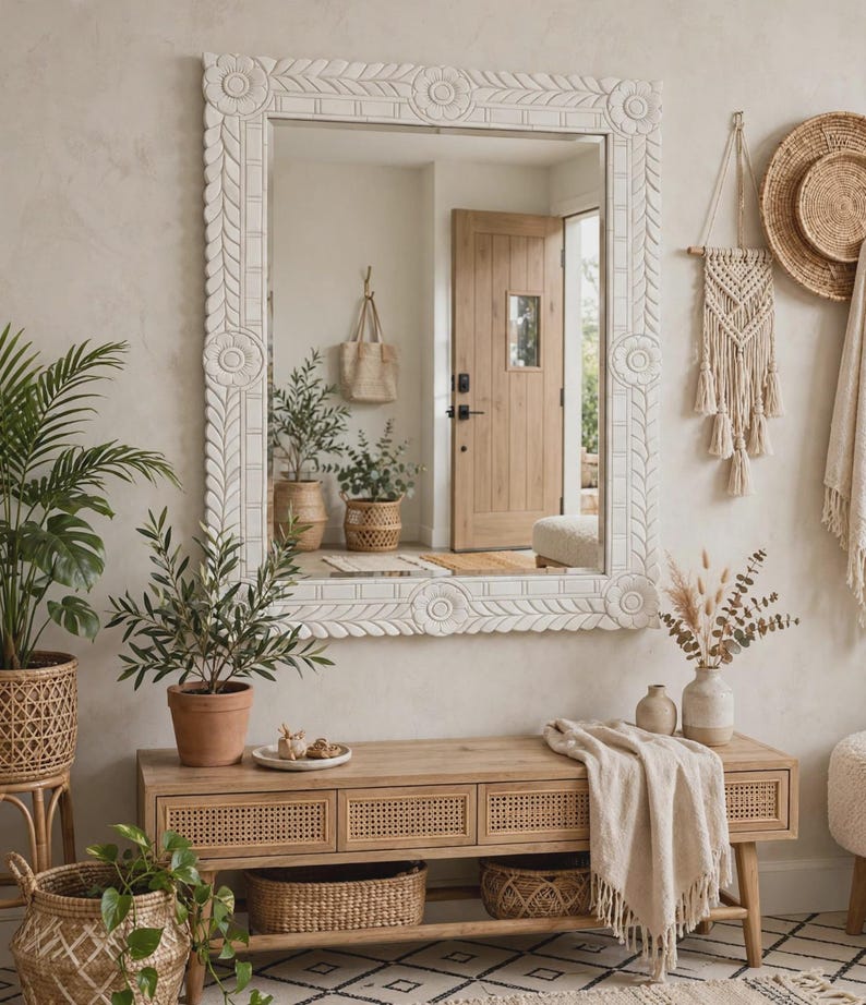 May include: A large rectangular wall mirror with a thick, off-white carved wooden frame featuring floral and leaf motifs. The mirror reflects an entryway with a wooden door and potted plants, while the frame has circular corner accents.