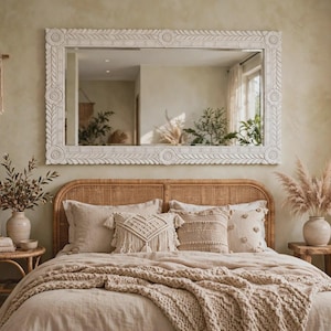 May include: A large rectangular wall mirror with a white carved wooden frame featuring floral and leaf motifs. The mirror is mounted above a bed with a natural rattan headboard, beige bedding, and several decorative throw pillows.