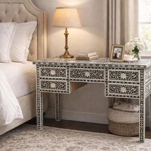 May include: A bedroom scene featuring a decorative desk with a floral pattern in black and white. The desk has multiple drawers and is topped with a lamp, books, and a framed photo. A bed with white bedding is visible.