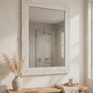 May include: A rectangular wall mirror featuring a white, hand-carved wooden frame with intricate floral motifs at each corner and a repeating leaf pattern along the edges. The mirror surface is clear and reflects a tiled shower area.