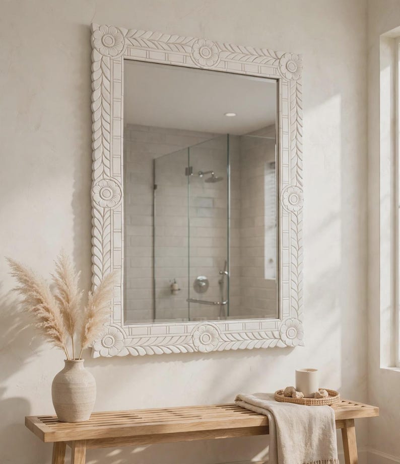 May include: A rectangular wall mirror featuring a white, hand-carved wooden frame with intricate floral motifs at each corner and a repeating leaf pattern along the edges. The mirror surface is clear and reflects a tiled shower area.