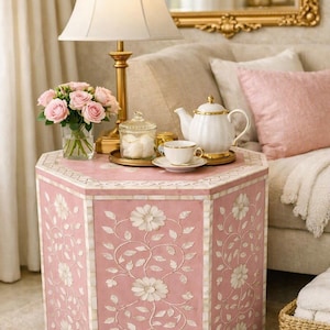 May include: A hexagonal side table featuring a pink surface with white floral and vine inlay patterns. The table has a light-colored border and a flat top, providing a stable base for a lamp, a glass vase with pink roses, and a tea set.