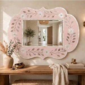 May include: A rectangular wall mirror with a scalloped frame in a soft pink hue, featuring an intricate inlay of white floral and leaf patterns. The reflective glass surface is centered within the decorative border, which has a smooth finish.