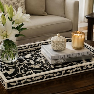 Puede incluir: Una bandeja rectangular en blanco y negro con un intrincado patrón floral incrustado. La bandeja contiene un jarrón de cristal transparente con lirios blancos, un pequeño recipiente con tapa y patrón geométrico, dos libros apilados titulados Live Beautiful e Ingrid, y una vela.