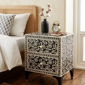 May include: A two-drawer bedside table featuring a black and white floral inlay pattern. The piece has a rectangular frame, two round knobs, and four dark legs with metallic feet. It is topped with a black vase containing cotton stems and a clock.