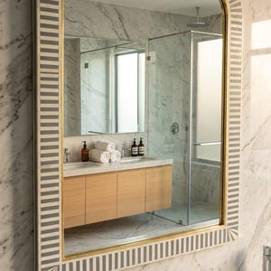 Puede incluir: Un espejo de baño grande y arqueado con marco dorado y borde decorativo de rayas grises y blancas. El espejo refleja un baño moderno con un mueble de baño de madera, paredes de mármol y una ducha de cristal.
