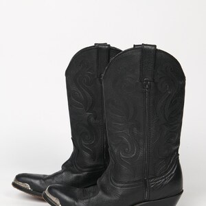 May include: A pair of black cowboy boots with decorative stitching and silver-toned metal accents on the toes. The boots have a classic Western design with angled heels and are set against a white background.