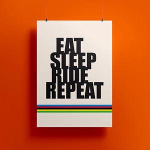 Op de afbeelding: Een witte poster met de woorden "EAT SLEEP RIDE REPEAT" in vette zwarte letters. Onder de tekst horizontale strepen in de kleuren van de Olympische ringen. De poster hangt tegen een oranje achtergrond.