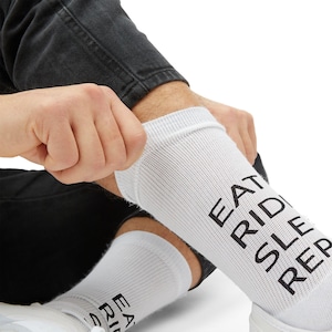 May include: White socks with the words "EAT RIDE SLEEP REPEAT" printed in black. The socks are being pulled up by a person wearing black jeans and white sneakers.