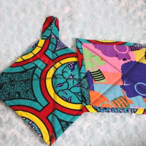 May include: Two colorful quilted pot holders. One is teal, red, yellow, and green with a circular pattern. The other is a patchwork of purple, pink, green, and blue with a silhouette design. The pot holders are on a light blue, textured surface.