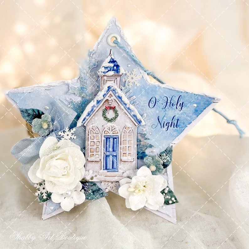 Op de afbeelding: Een blauwe en witte stervormige kerstversiering met een witte kerk en de tekst "O Holy Night". De versiering is versierd met witte bloemen, blauw lint en sneeuwvlokken.