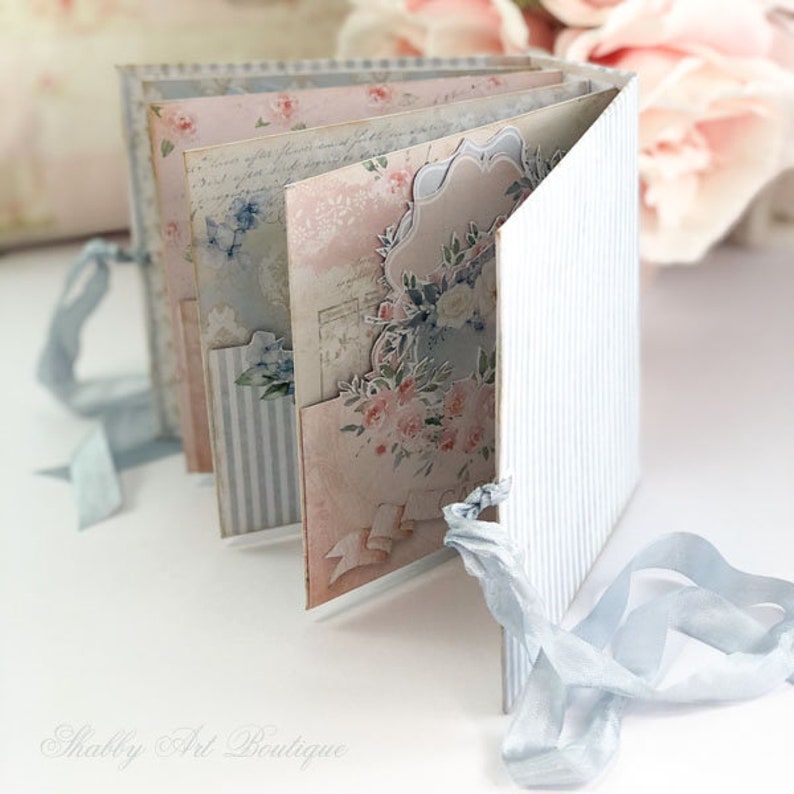 May include: A small, handmade scrapbook with a blue and white striped cover and a floral design. The scrapbook is closed and tied with a blue ribbon.