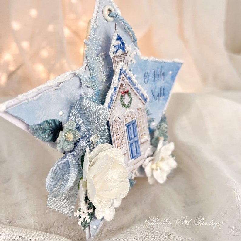 Op de afbeelding: Een blauwe en witte stervormige kerstversiering met een witte kerk, een blauw lint en de tekst "O Holy Night".