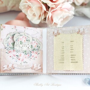 May include: A pink and blue scrapbook with a lace doily and floral design. The scrapbook has a pocket for thread and a section for needles.