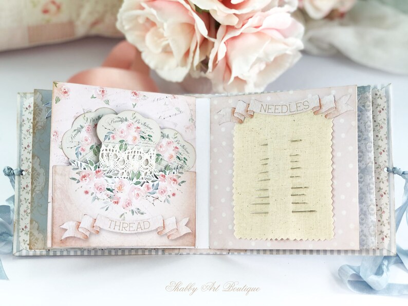 May include: A pink and blue scrapbook with a lace doily and floral design. The scrapbook has a pocket for thread and a section for needles.