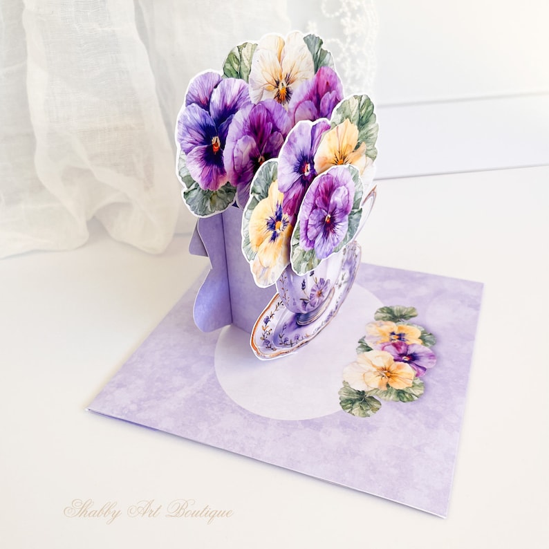 Peut inclure: Une carte pop-up violette et blanche avec une tasse &agrave; th&eacute; remplie de pens&eacute;es violettes et jaunes. La carte a un fond violet avec un cercle blanc au centre et le texte "Shabby Art Boutique" en bas.