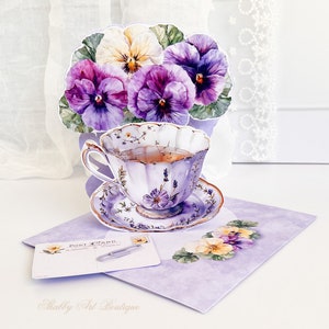 Peut inclure: Une tasse &agrave; th&eacute; et une soucoupe violettes et blanches avec un motif floral. La tasse &agrave; th&eacute; est remplie de th&eacute; et est entour&eacute;e d'un bouquet de pens&eacute;es violettes et blanches. La tasse &agrave; th&eacute; et la soucoupe sont sur un fond violet avec le texte "Postcard" et "Shabby Art Boutique".