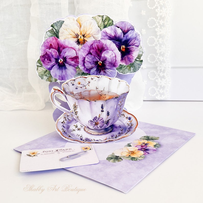 Peut inclure: Une tasse &agrave; th&eacute; et une soucoupe violettes et blanches avec un motif floral. La tasse &agrave; th&eacute; est remplie de th&eacute; et est entour&eacute;e d'un bouquet de pens&eacute;es violettes et blanches. La tasse &agrave; th&eacute; et la soucoupe sont sur un fond violet avec le texte "Postcard" et "Shabby Art Boutique".