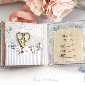 May include: A small, blue and white fabric-covered book with a pocket on the left side holding a pair of gold scissors. The right side of the book has a piece of beige fabric with a row of safety pins labeled "SAFETY PINS".