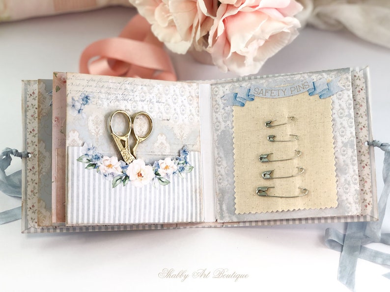 May include: A small, blue and white fabric-covered book with a pocket on the left side holding a pair of gold scissors. The right side of the book has a piece of beige fabric with a row of safety pins labeled "SAFETY PINS".