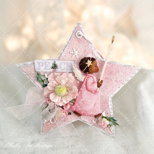 Op de afbeelding: Een roze stervormige ornament met een witte engel die een kaars vasthoudt. De engel heeft een bruine huid en zwart haar. De ornament is versierd met een roze bloem, een groene hulsttak en het woord "Vrede" in witte letters.