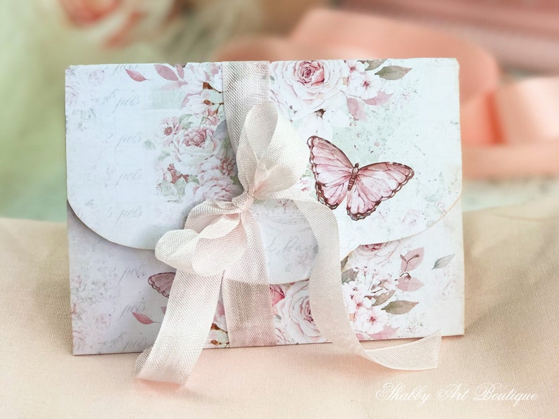 May include: A white envelope with a floral design and a pink butterfly. The envelope is tied with a white ribbon and has the text "Shabby Art Boutique" printed on the bottom.