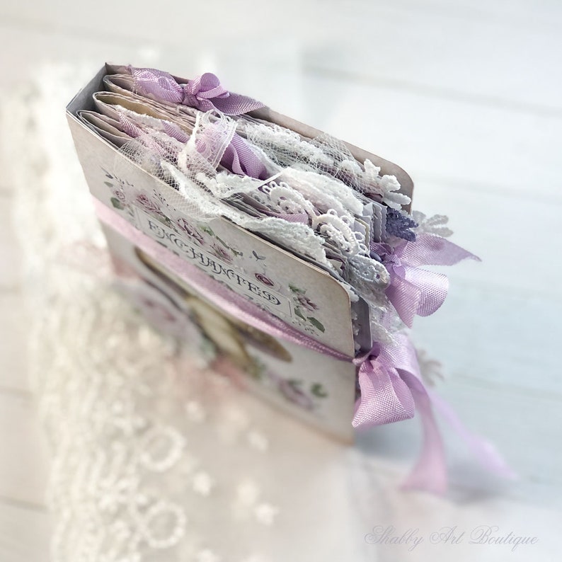 May include: A lavender and white journal with lace and ribbon embellishments. The cover features the text "Forget Me Not" in a floral design.
