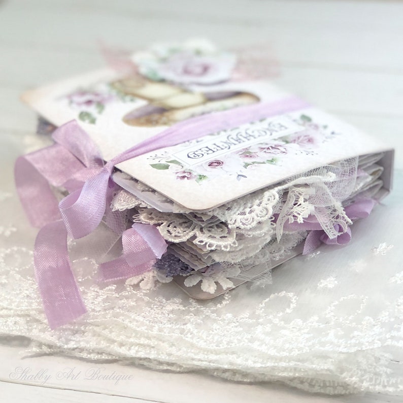 May include: A small, handmade book with a white cover and purple ribbon. The cover features a floral design and the text "The Memories". The book is decorated with lace and is sitting on a white lace surface.