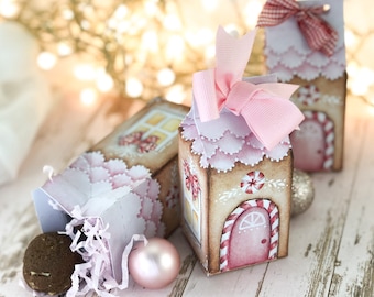 May include: Three small gingerbread house-shaped gift boxes with pink and white candy cane details. The boxes are decorated with pink and white bows and have a white background with pink accents. The boxes are sitting on a white wooden surface.