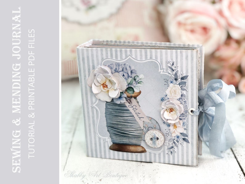 May include: A blue and white striped journal with a floral design and a spool of thread on the cover. The journal is tied with a blue ribbon and has the text "Sewing & Mending Journal" on the front.